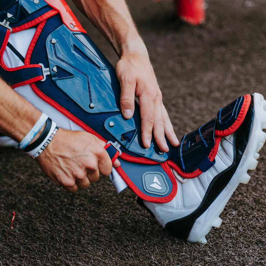 An adult fastens All Star MVP Pro Catcher's Leg Guards: LG-5, featuring a pivoting hinge system, over white cleats while preparing for a game on the turf field.