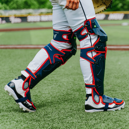 A catcher wearing white pants and All Star MVP Pro Catcher's Leg Guards: LG-5 in navy with red accents stands on artificial turf. The All Star gear features a pivoting hinge system for mobility; only the lower body is visible.