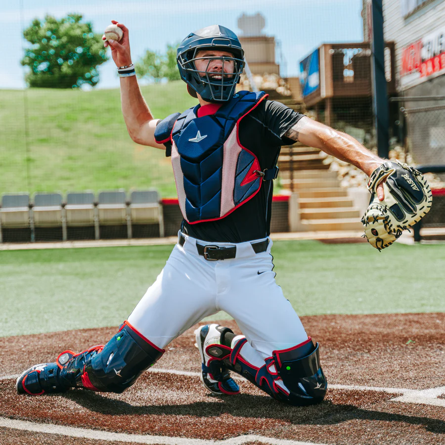 A baseball catcher, wearing an All Star MVP Pro Catcher's Chest Protector (CPCC-5), kneels on the field, ready to throw a ball with his right hand while holding a catcher’s mitt in his left.