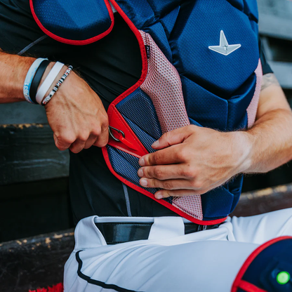 A baseball catcher tightens the strap on an All Star MVP Pro Catcher's Chest Protector (CPCC-5), featuring blue and red panels, while sitting on a bench in white pants and wearing several wristbands.