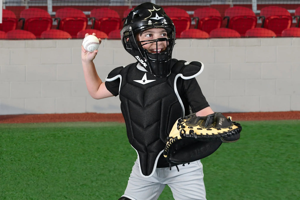 A young starting catcher, outfitted in the All Star Future Star Catcher’s Chest Protector: CP-FS by All Star and full protective gear including shoulder caps, stands ready on a green field with red stadium seats in the background, holding a ball in one hand and a glove in the other.