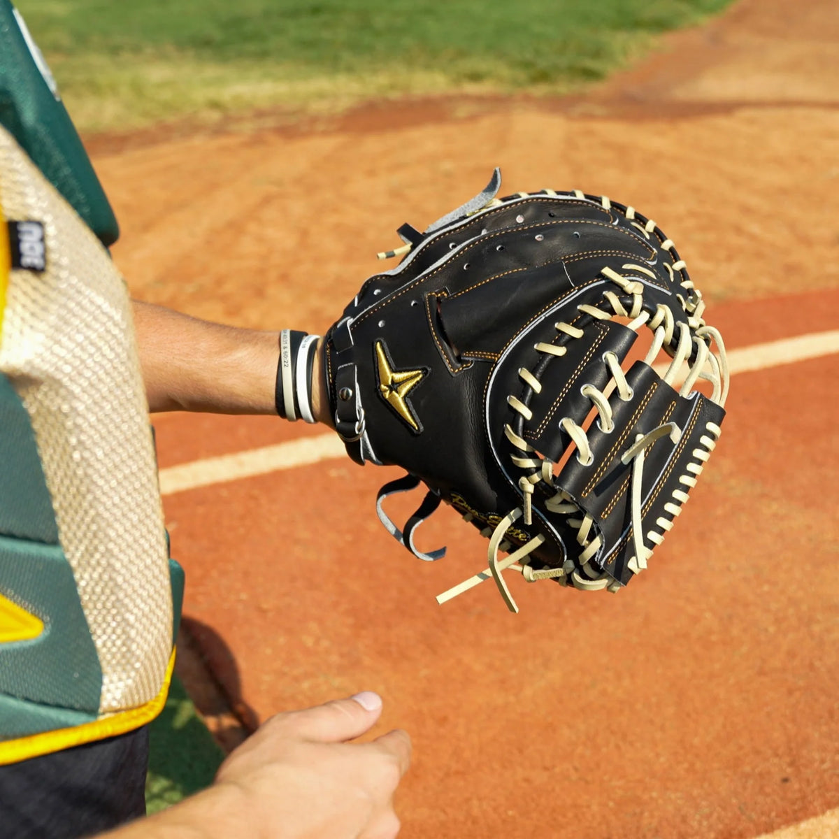 A person stands on a dirt field, ready to catch a ball while wearing the All Star Pro-Elite Japan 34″ Baseball Catcher’s Mitt: CM3000-J1-34 by All Star. The mitt is crafted from premium HIMEJI leather, showcasing black material with white laces. In the background, grass and a white foul line can be seen.