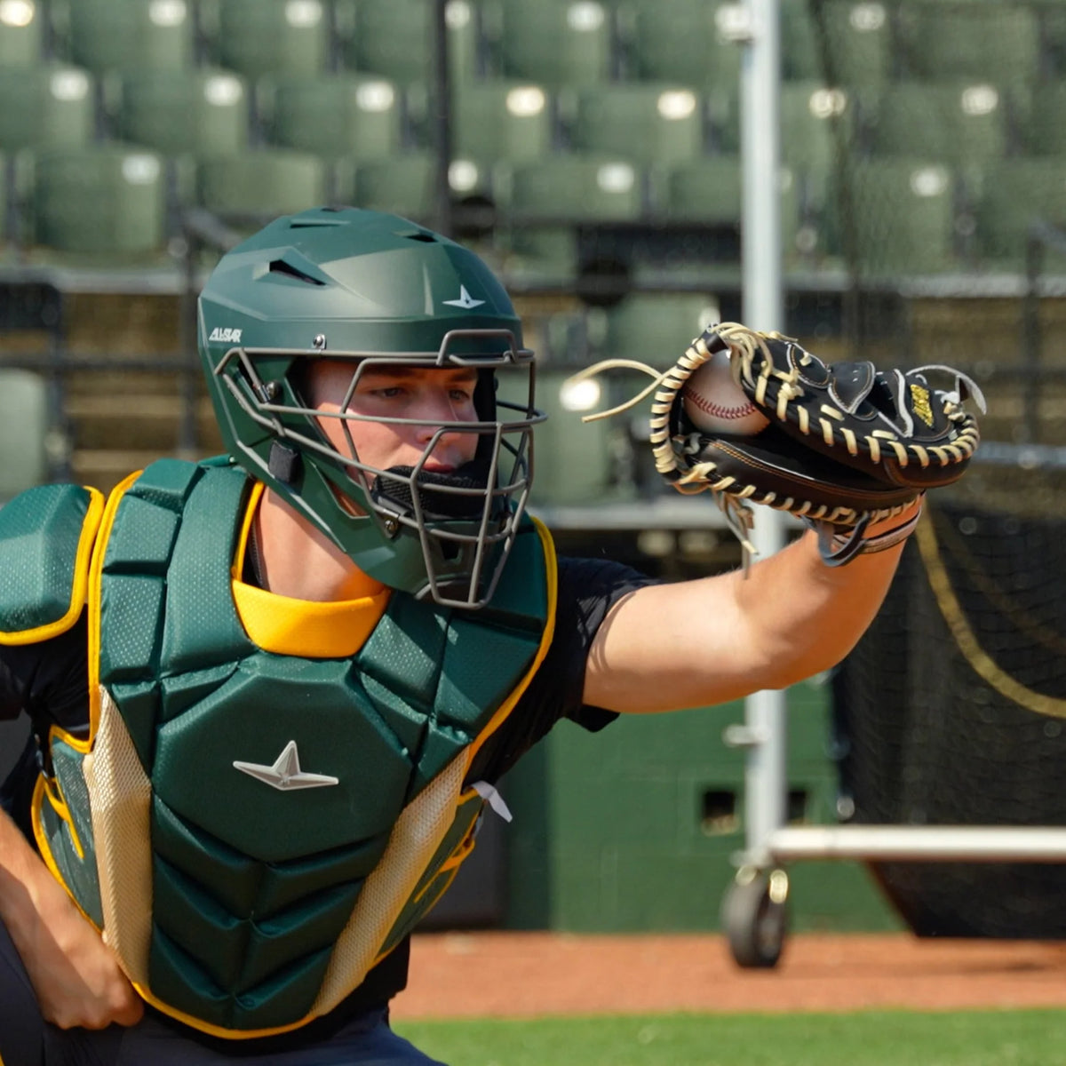 A baseball catcher in green and yellow gear reaches out with an All Star Pro-Elite Japan 34″ Baseball Catcher’s Mitt (CM3000-J1-34) to catch a baseball during a game, with empty stadium seats in the background, evoking scenes from the Japan Series.