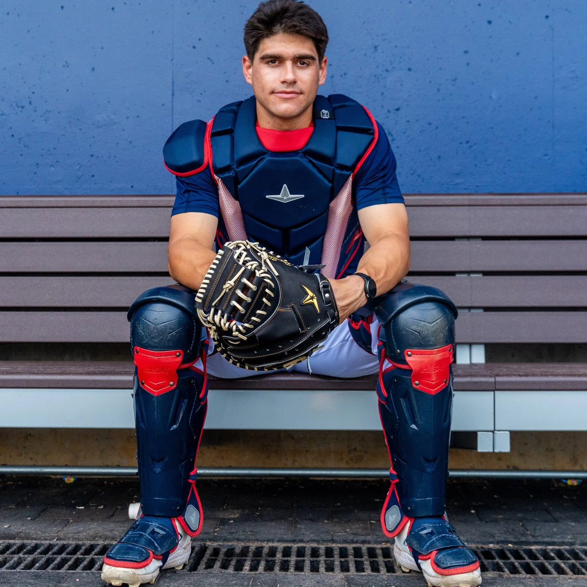 An All Star Pro-Elite Japan 34″ Baseball Catcher’s Mitt (CM3000-J1-34), expertly crafted from premium HIMEJI leather, is held by a baseball catcher in full protective gear as he sits on a bench and looks directly at the camera against a blue background.