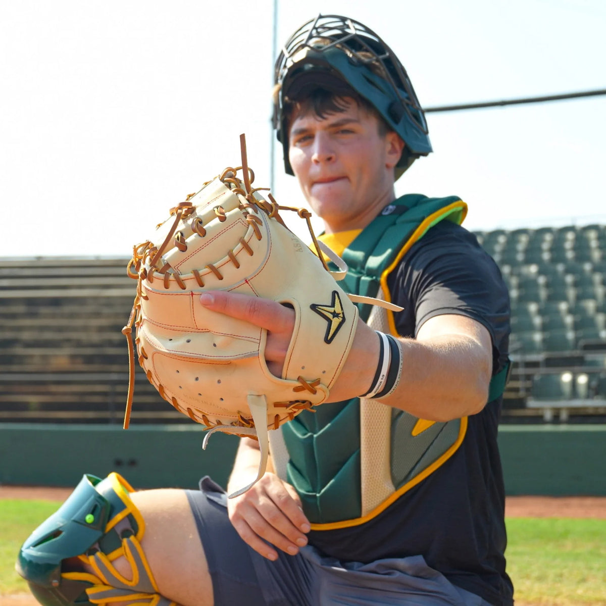 Wearing full catching gear, a baseball catcher stands on a field with empty bleachers in the background, holding up the All Star Pro-Elite Japan 34″ Baseball Catcher’s Mitt (CM3000-J2-34) by All Star toward the camera.