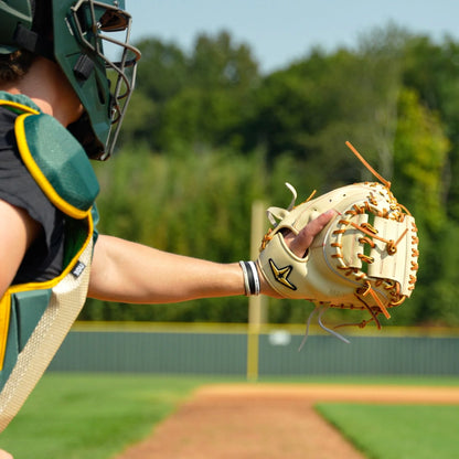 Wearing full gear, a catcher stands ready on a sunny field, extending their All Star Pro-Elite Japan 34″ Baseball Catcher’s Mitt: CM3000-J2-34 to catch the ball. The vibrant green background features trees and a fence.