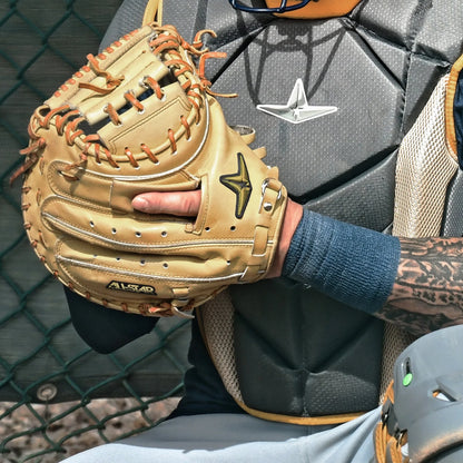 An All Star Pro-Elite Japan 34″ Catcher’s Mitt (CM3000-J2-34), expertly crafted from premium HIMEJI leather, is held close to the chest by a catcher in full protective gear standing beside a chain-link fence.
