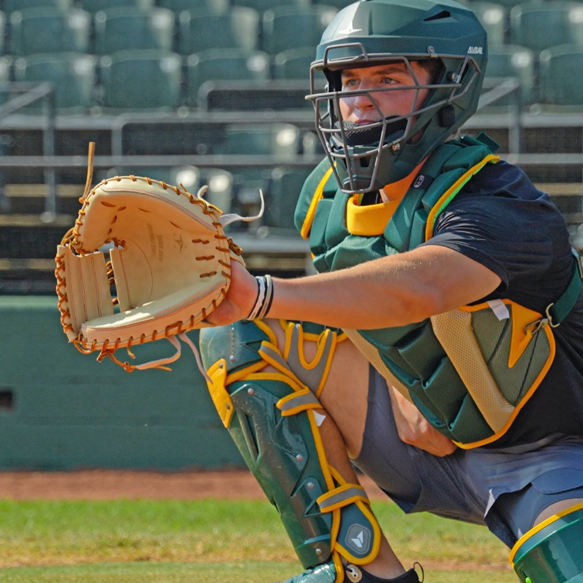 A baseball catcher in full gear crouches behind home plate with the All Star Pro-Elite Japan 34″ Baseball Catcher’s Mitt (CM3000-J2-34), crafted from premium HIMEJI leather, ready to catch a pitch. In the background, empty stadium seats and a railing are visible.