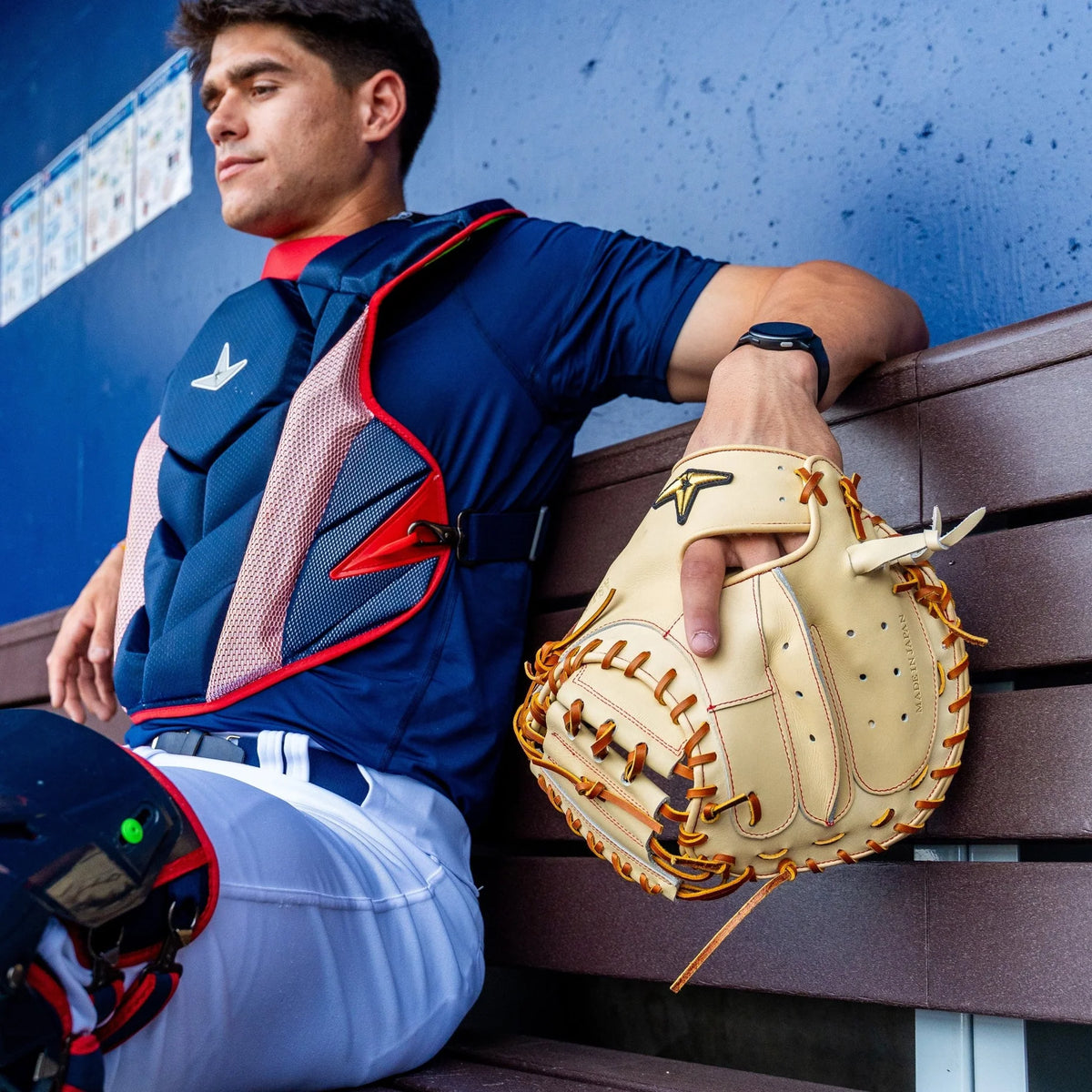 A baseball catcher in protective gear sits on a bench, holding an All Star Pro-Elite Japan 34″ Catcher’s Mitt (CM3000-J2-34), expertly crafted from premium HIMEJI leather. He wears a navy shirt, white pants, and a chest protector, looking off to the side in the dugout.