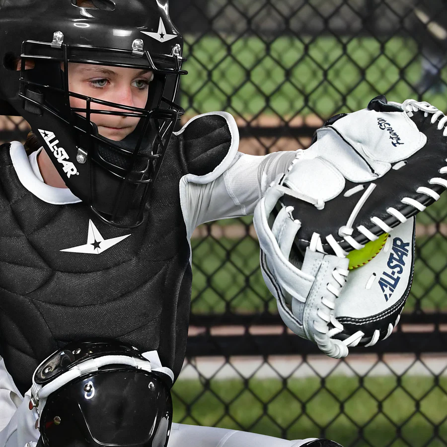 A young softball catcher in full gear squats behind home plate, holding an All Star Future Star 32.5" Fastpitch Catcher's Mitt (CMW-FS-Y) with a yellow softball inside and an adjustable wrist strap; a chain-link fence is visible behind.