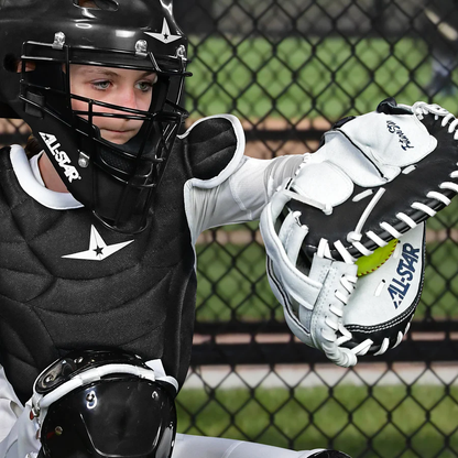 A young softball catcher in full gear squats behind home plate, holding an All Star Future Star 32.5" Fastpitch Catcher's Mitt (CMW-FS-Y) with a yellow softball inside and an adjustable wrist strap; a chain-link fence is visible behind.