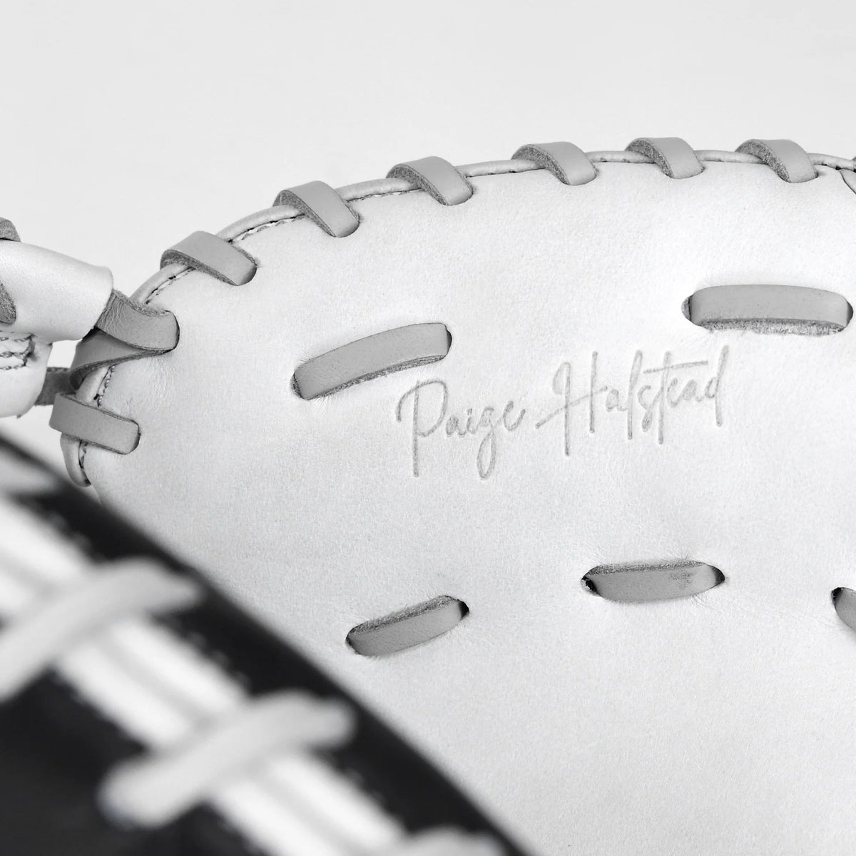 A close-up of the All Star PHX 33″ Paige Halstead Fastpitch Catcher’s Mitt (CMW-PHX-33) showcases its white leather construction with gray laces and “Paige Halstead” embossed on the glove, featuring an adjustable pocket system designed by All Star for a perfect travel ball fit.