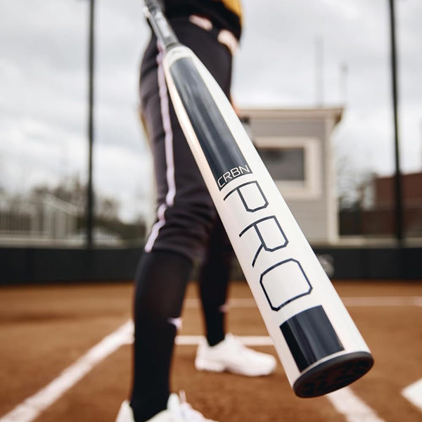 A person in athletic clothing stands on a baseball field, holding a white bat labeled PRO toward the camera, creating a dramatic close-up perspective. The background shows part of the field and a building.