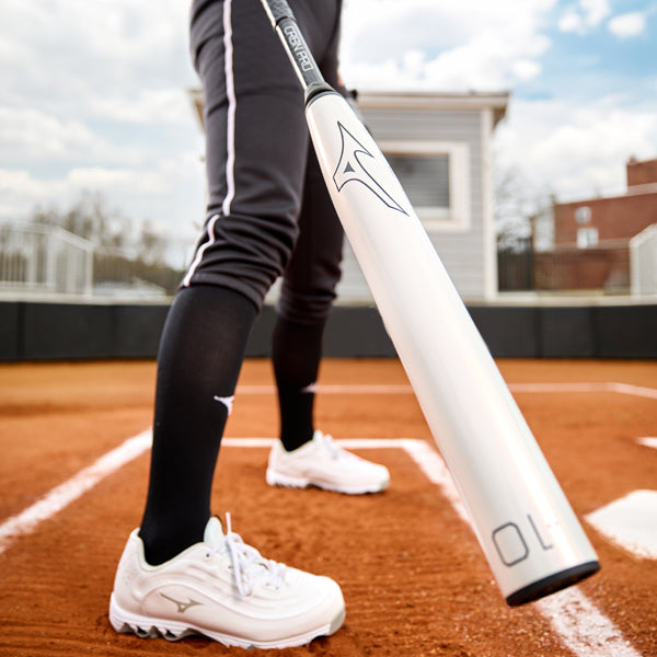 A person in black athletic pants and white shoes stands on a baseball field, holding a silver baseball bat over home plate. The focus is on the bat and their lower body.