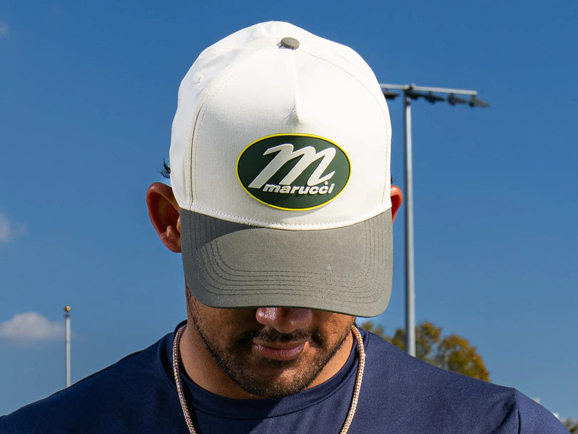 A man wearing a white and gray Marucci baseball cap with a large M logo faces downward outdoors under a clear blue sky.