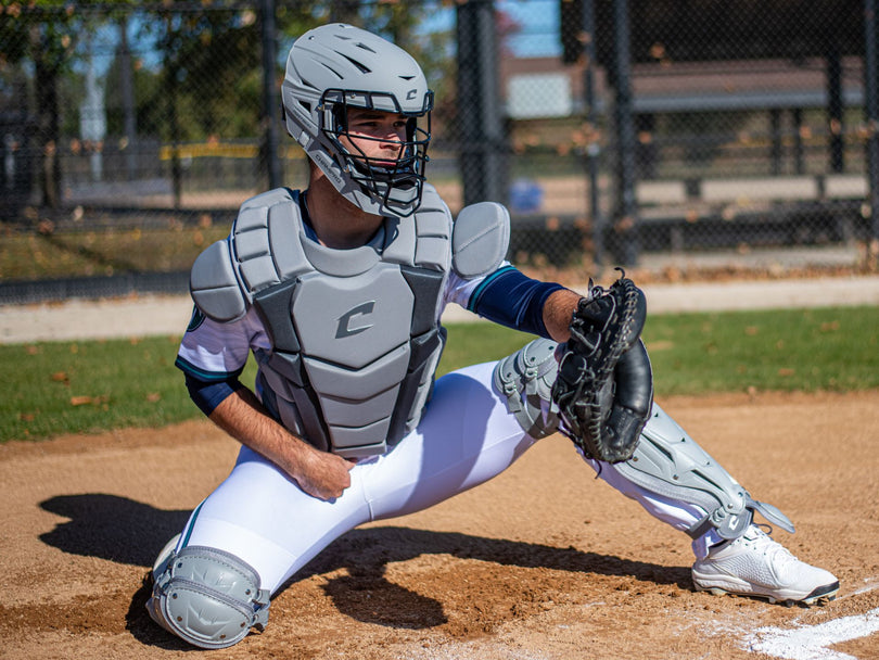 A baseball catcher wearing full gray protective gear and a helmet crouches on a dirt field, glove extended to catch a ball, with a chain-link fence and trees in the background.