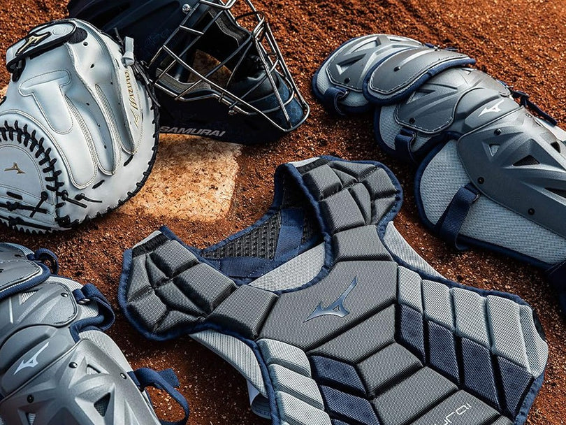 Baseball catcher’s gear, including a mitt, helmet, chest protector, and leg guards, is laid out on a dirt field. The equipment is mostly gray with navy blue and white accents.