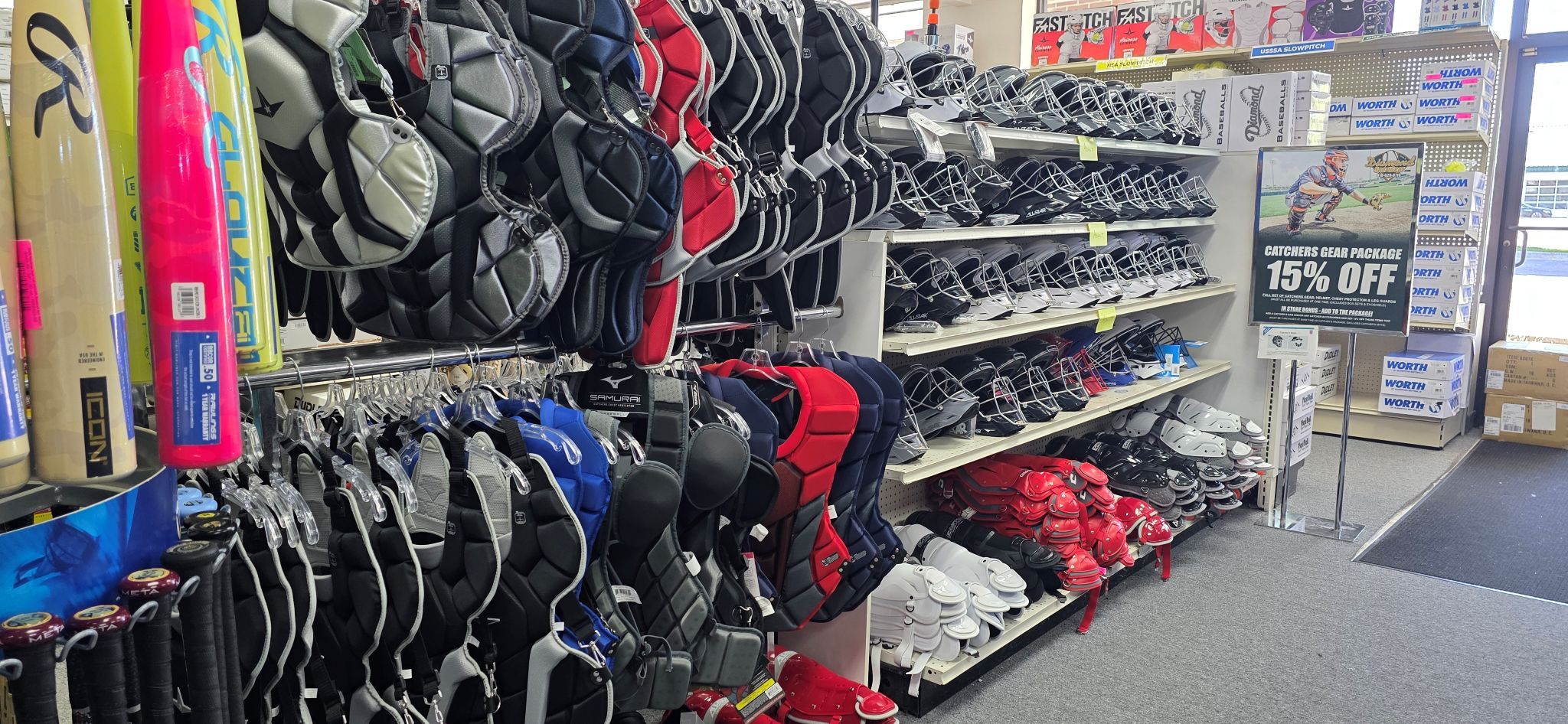 Rows of baseball and softball equipment in a sporting goods store, including bats, catchers chest protectors, helmets, and cleats. A sign advertises a 15% off sale near the entrance.