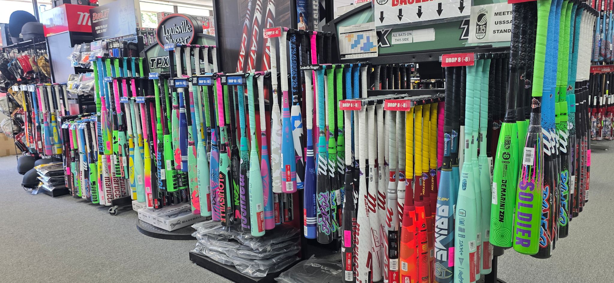 Rows of colorful baseball and softball bats are displayed on racks in a sporting goods store, with various brands and models visible. Accessories and gear are also seen in the background.