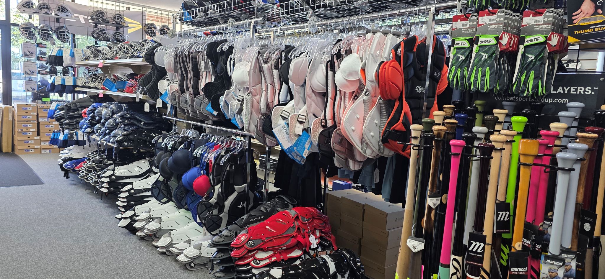 A sporting goods store display with shelves of protective gear, including helmets and catching masks, racks of gloves, and bins of colorful baseball and softball bats. Shoes and boxes are visible on the left side.
