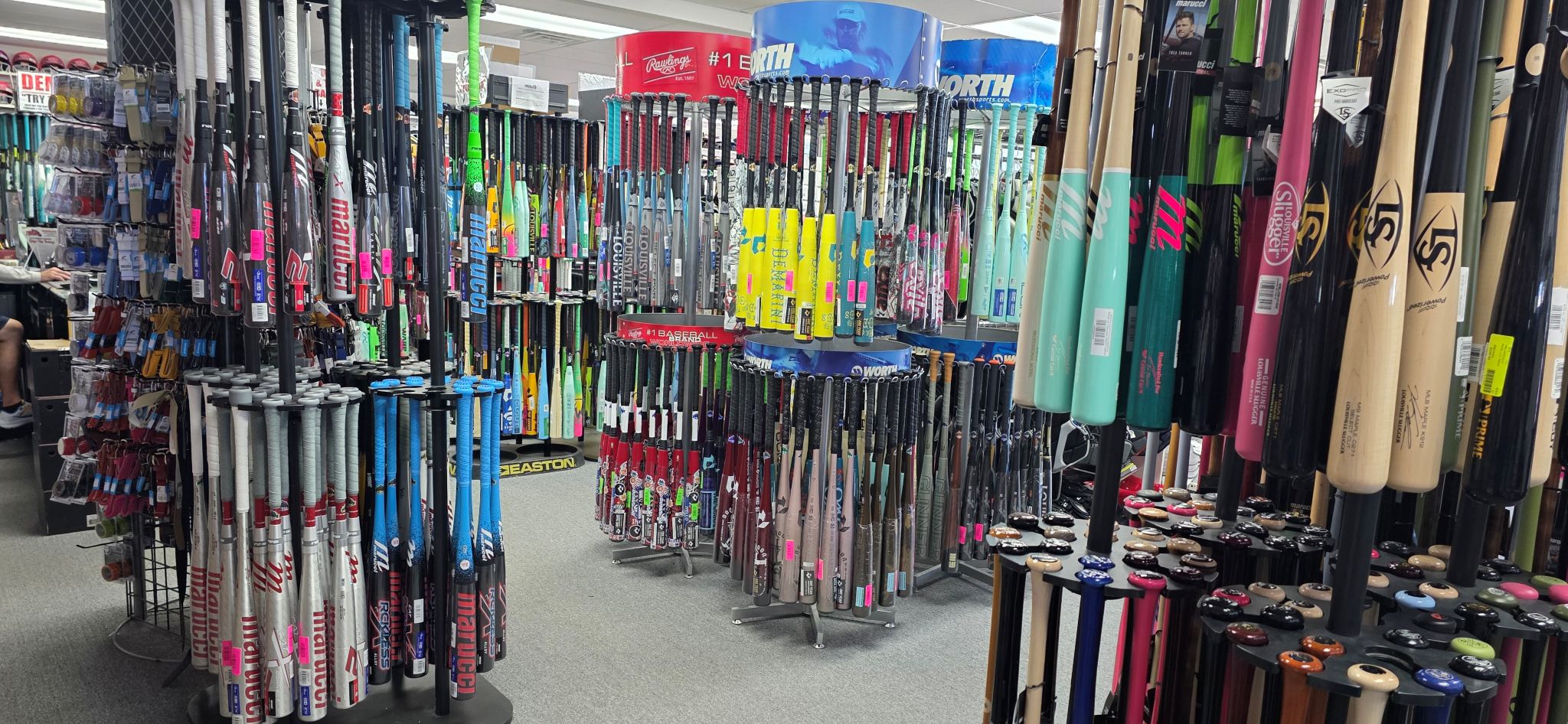 A sporting goods store with numerous baseball and softball bats displayed vertically on racks, featuring various colors and brands. The scene shows a dense selection with bats arranged closely together.