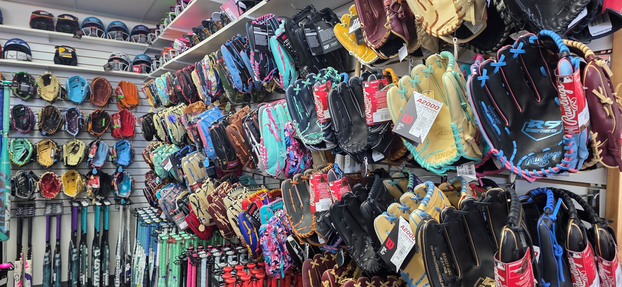 Rows of baseball and softball gloves in various colors and brands are displayed on wall racks, with multiple bats and helmets visible along the left side of the store.