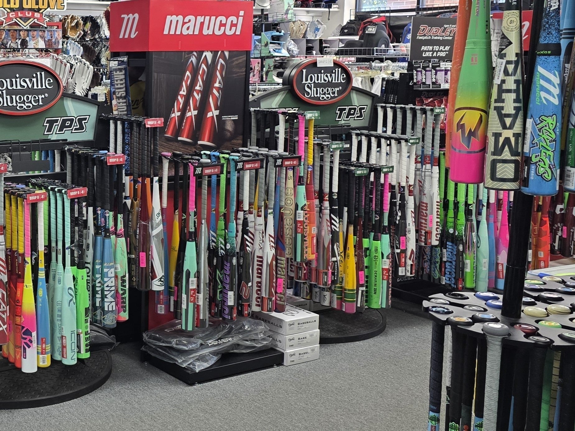 A sporting goods store display showcases a wide variety of colorful baseball and softball bats from brands like Louisville Slugger and Marucci, arranged upright in racks with sports gear visible in the background.