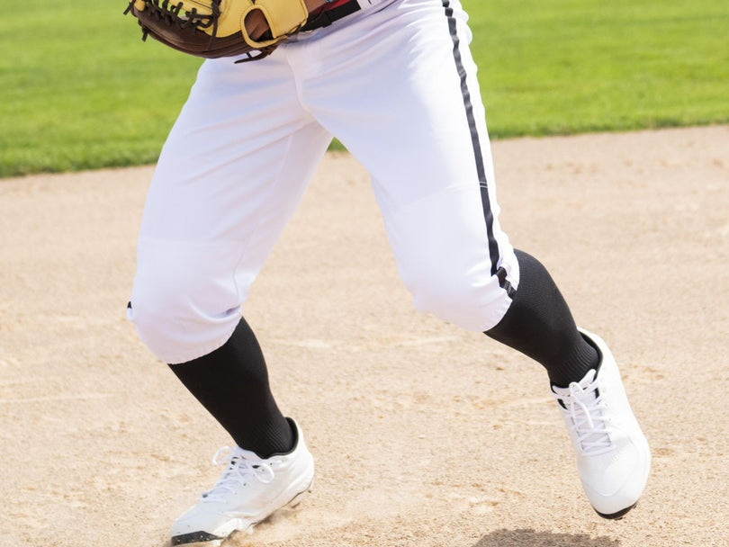 A baseball player in white pants, black socks, and white cleats runs on a baseball field with a glove on their left hand. The image is cropped to show only the lower body.