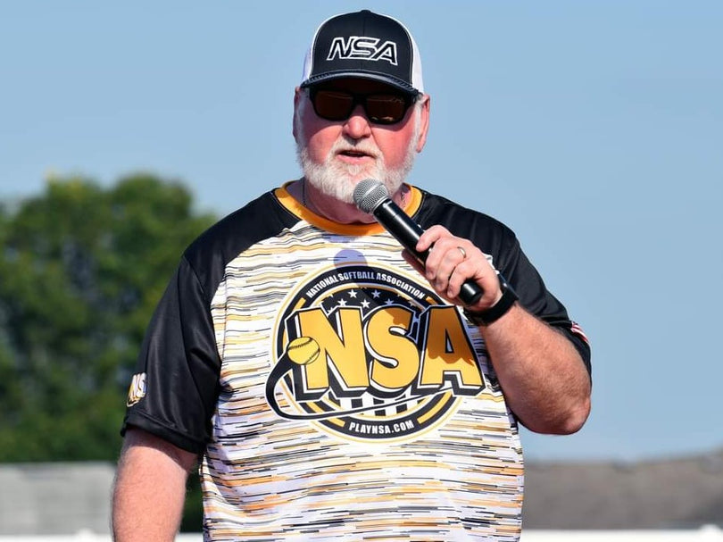 A man wearing sunglasses, a black-and-white NSA cap, and an NSA sports jersey speaks into a microphone outdoors under a clear sky.