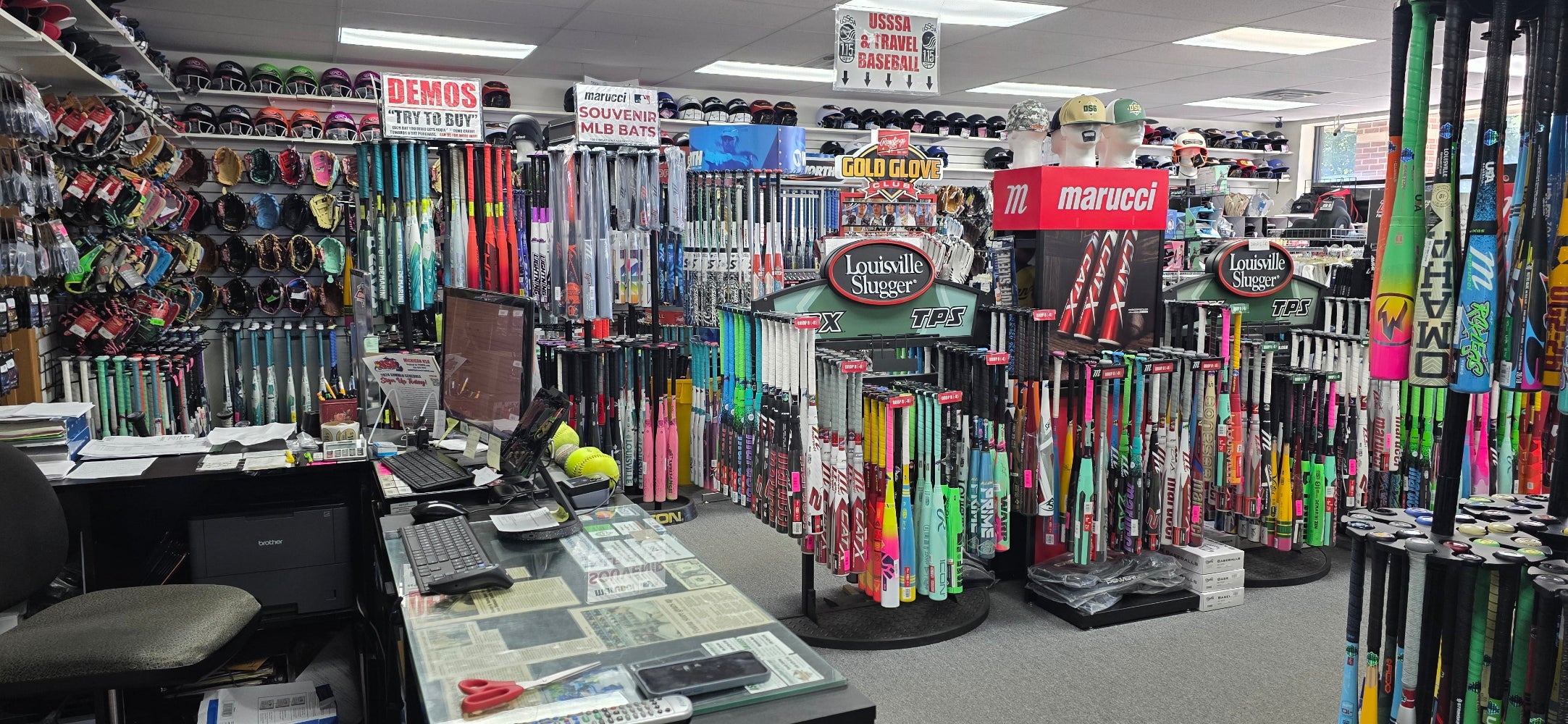 A sporting goods store interior filled with colorful baseball bats, gloves on wall racks, mannequins wearing hats, and a cluttered front counter with a computer, phone, and newspapers. Signs and displays promote various brands.