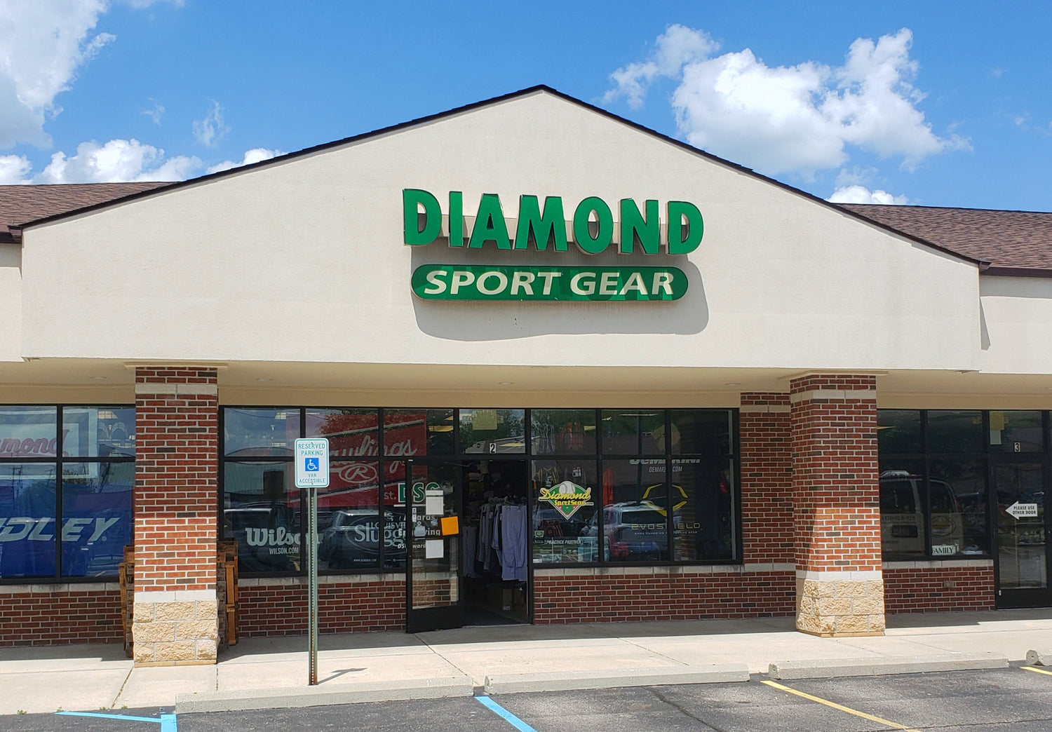 Storefront of DIAMOND SPORT GEAR with a green sign above the entrance, brick columns, large windows, and a partly cloudy sky overhead. A handicapped parking sign is visible in front.