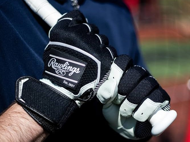 A person wearing black and white Rawlings batting gloves grips a white baseball bat, with a blurred field in the background.