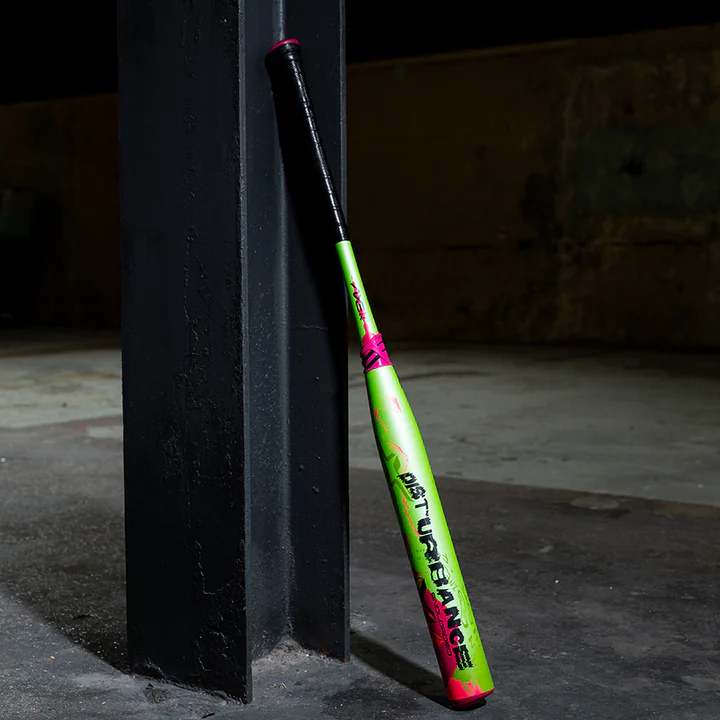 A green and pink baseball bat with black tape on the handle leans against a dark metal pillar in an empty industrial space with a concrete floor.