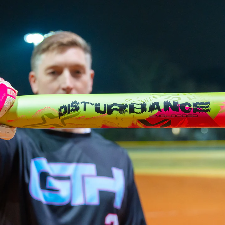 A man holds a neon yellow and pink baseball bat labeled DISTURBANCE toward the camera, with a blurred baseball field and night lights in the background.