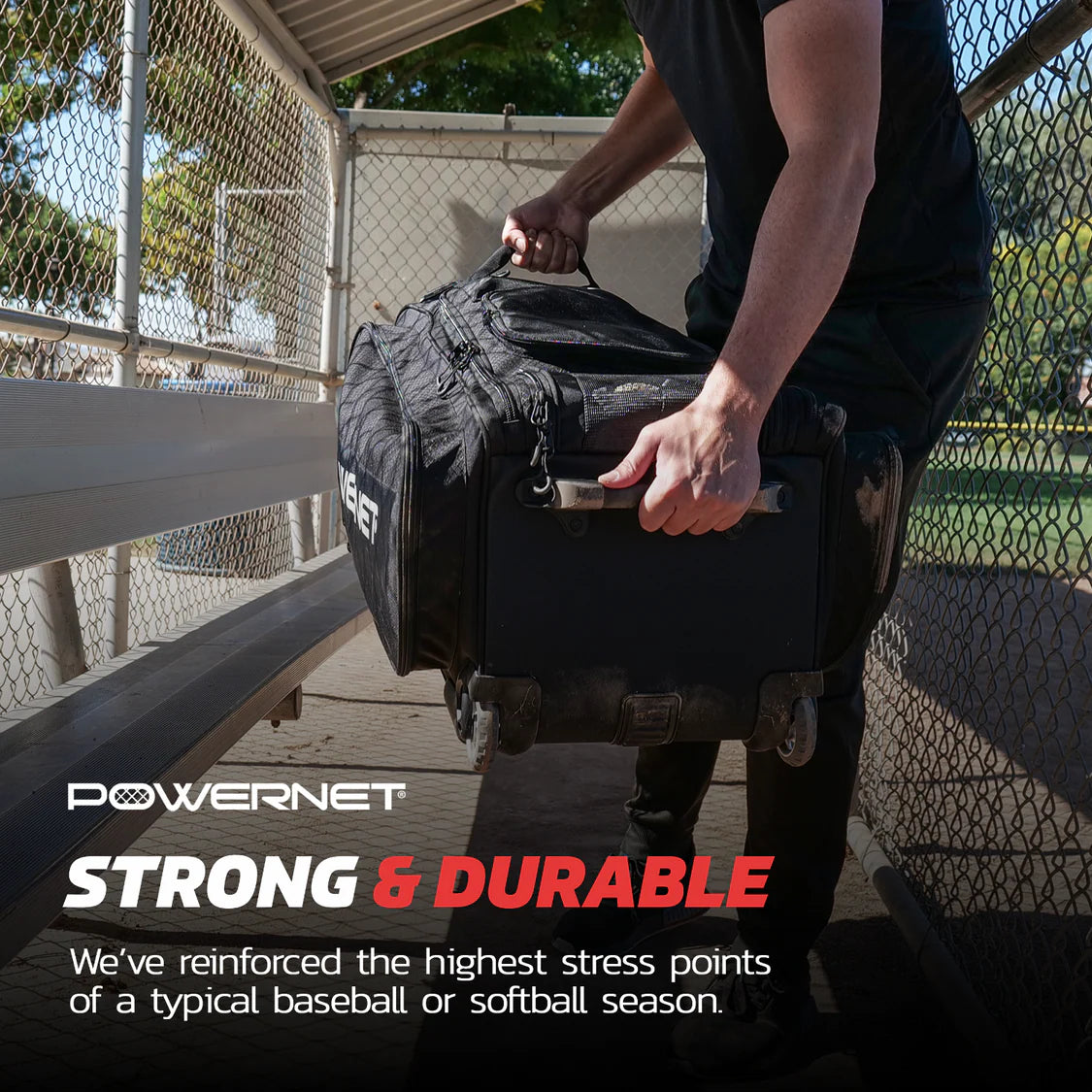 A person lifts the PowerNet Optimus Catcher's Gear Bag: B013-BK, a large black rolling bag with wheels and handles, inside a chain-link dugout. The PowerNet bag is strong and durable with reinforced stress points—ideal for baseball or softball seasons.