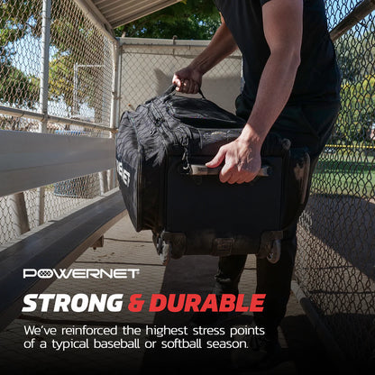 A person lifts the PowerNet Optimus Catcher's Gear Bag: B013-BK, a large black rolling bag with wheels and handles, inside a chain-link dugout. The PowerNet bag is strong and durable with reinforced stress points—ideal for baseball or softball seasons.