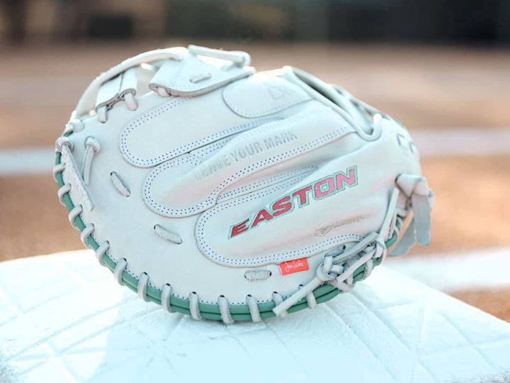 A close-up of a white Easton baseball glove with teal stitching and the phrase LEAVE YOUR MARK embossed on it, resting on a white surface, with a blurred background.