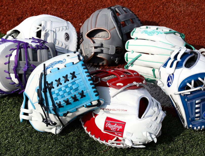 A close-up of six colorful softball gloves, each with unique designs and laces, arranged on artificial turf. The gloves feature colors like white, blue, red, purple, gray, and green.