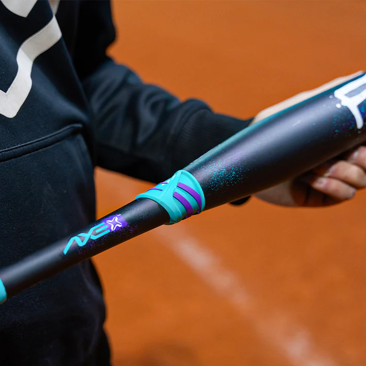 A person wearing a dark hoodie holds a black baseball bat with blue and purple accents over a reddish-brown field.
