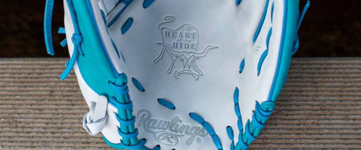 A close-up of a white and blue Rawlings baseball glove, showing the Heart of the Hide logo and stitching details inside the pocket, resting on a wooden surface.
