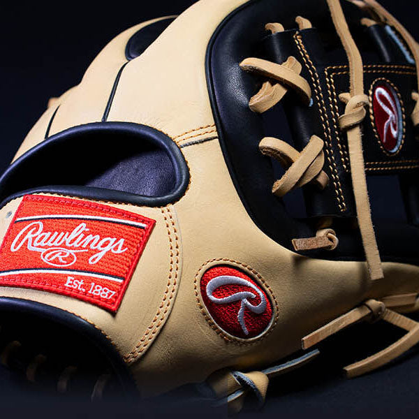 Close-up of a tan and black Rawlings baseball glove, showing detailed stitching, red Rawlings logo patches, and tan leather laces against a dark background.
