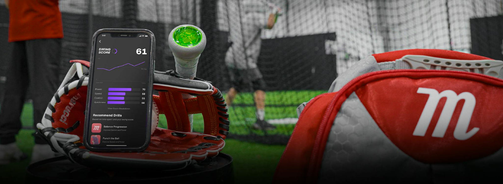 A smartphone displaying baseball analytics rests on a red baseball glove beside a bat and a catchers mitt with an M logo. In the blurred background, people practice baseball in an indoor batting cage.