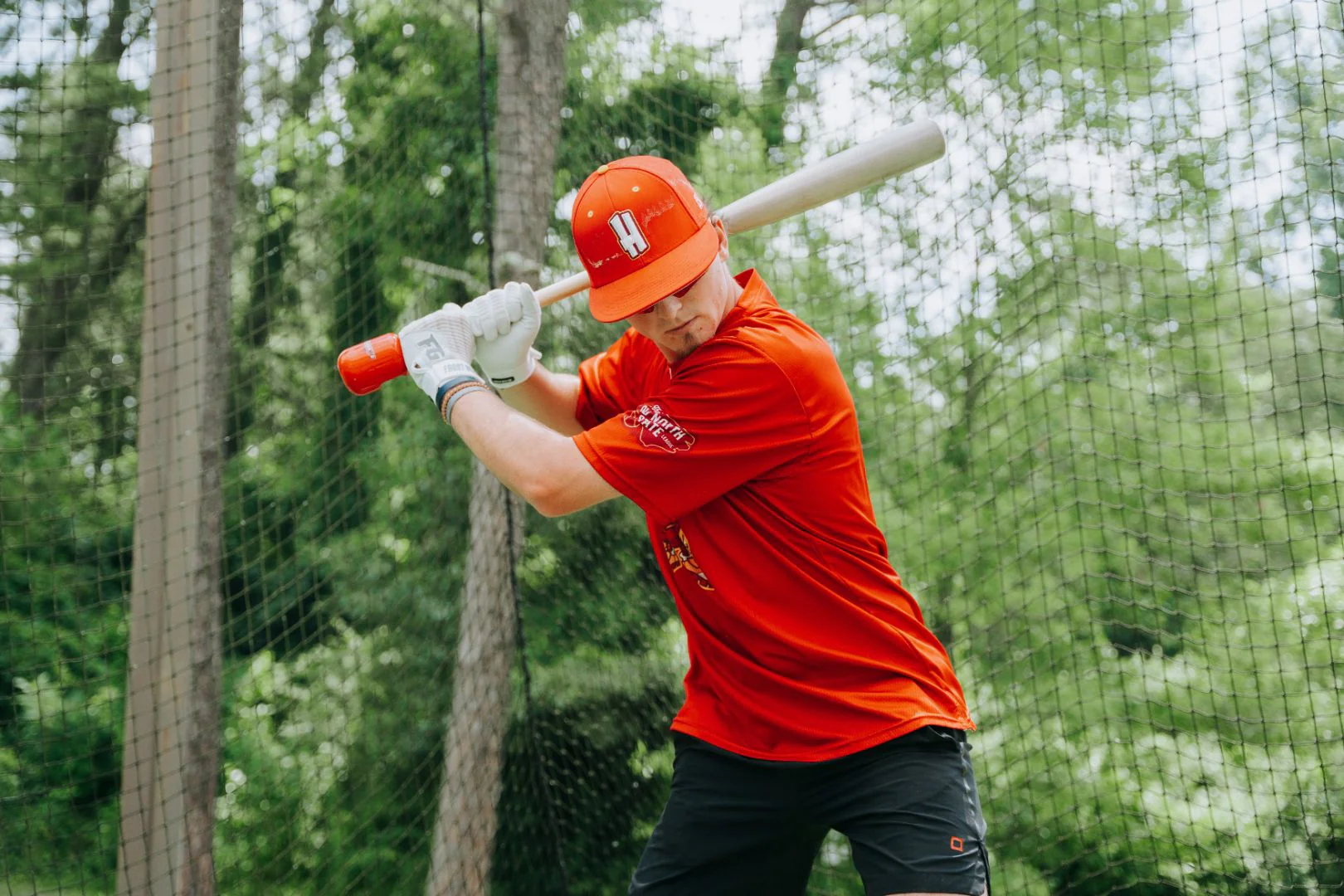 A baseball player in a red jersey and helmet stands ready to bat, holding a bat over his shoulder, with trees and a netted fence in the background.