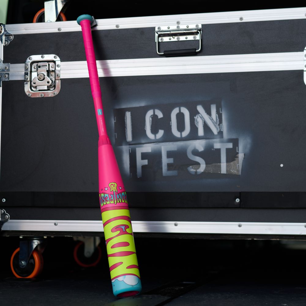 A vibrant limited edition Rawlings 2026 Icon Fest (-10) 2 5/8" USA Baseball Bat (RUS6IFP10) with pink, green, and blue designs leans against a black equipment case labeled ICON FEST in white stencil.