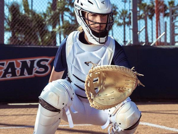 A softball catcher wearing a helmet, chest protector, and leg guards crouches behind home plate with a mitt ready, on a sunny field with palm trees and a fence in the background.
