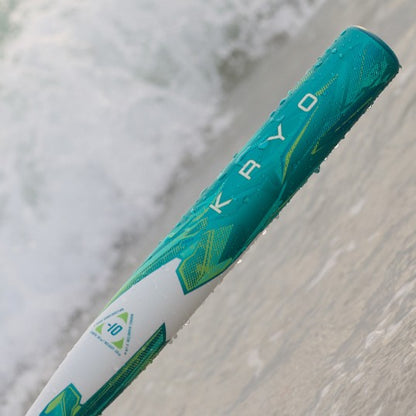 The 2026 Louisville Slugger Kryo (-8) Fastpitch Softball Bat (WBL4137010), with teal and white LOUISVILLE SLUGGER branding, rests on wet sand by the shore as ocean waves approach in the background.