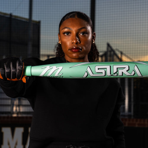 A woman in black sportswear holds a mint green Marucci 2025 ASURA (-9) Fastpitch Softball Bat (MFPAS9 DEMO) with Gap Sync barrel technology horizontally toward the camera on an outdoor field with a fence and buildings behind her.