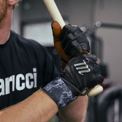 A person wearing Marucci REMX Youth Batting Gloves (MBRMXY) grips a wooden bat. Dressed in a black shirt with white text, they stand in an indoor sports facility.
