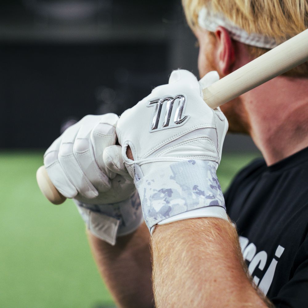 A youth with light hair in a black shirt grips a bat, preparing to swing indoors, while wearing Marucci REMX Youth Batting Gloves (MBRMXY) featuring the silver Marucci logo.
