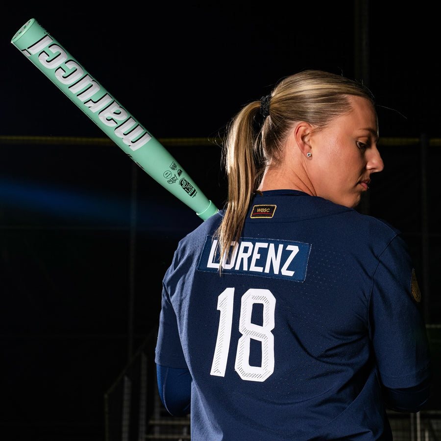 A softball player in a navy jersey with LORENZ and number 18 on the back holds a 2025 Marucci ASURA (-9) Fastpitch Softball Bat: MFPAS9 (DEMO), highlighting its Gap Sync barrel tech against a dark background.